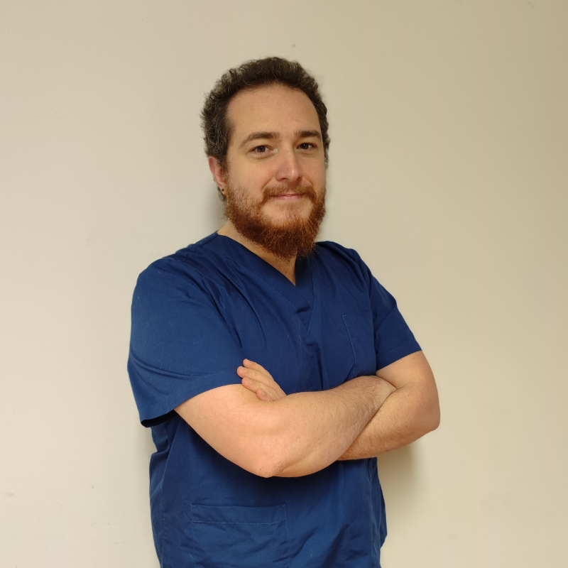 Una persona con barba e capelli ricci, vestita con un camice da medico blu, è in piedi contro una semplice parete chiara con le braccia incrociate e un leggero sorriso.