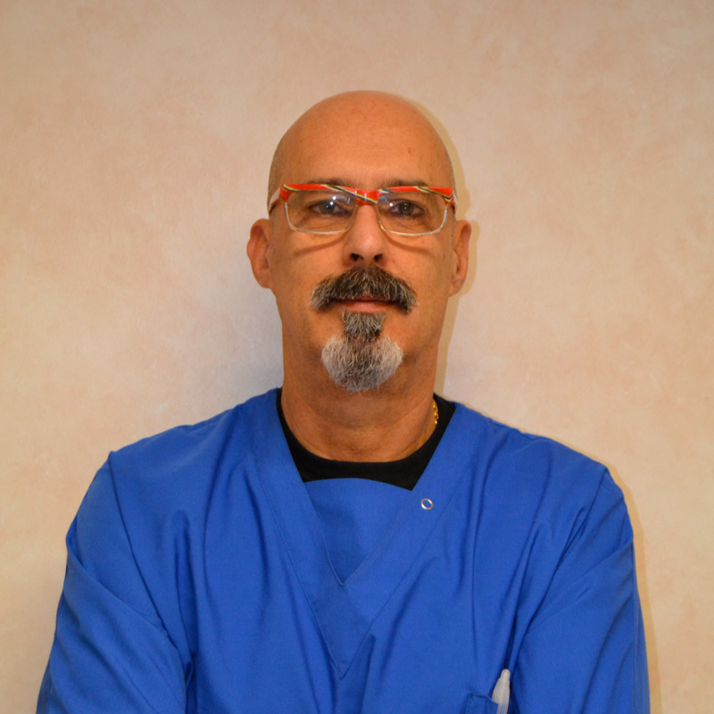 Un uomo con testa calva, baffi e pizzetto indossa occhiali con montatura rossa e un camice da medico blu, in piedi su uno sfondo beige con le braccia incrociate.