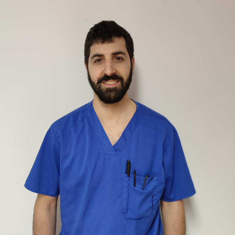 Un uomo con i capelli corti e scuri e la barba sorride, indossa un camice da medico blu con delle penne nel taschino, in piedi contro una semplice parete chiara.