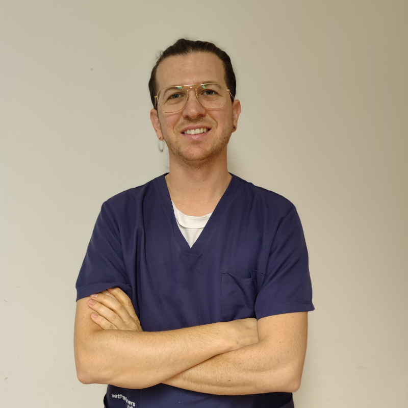 Una persona che indossa occhiali e camice blu sta in piedi contro una parete chiara, sorridendo con le braccia incrociate.