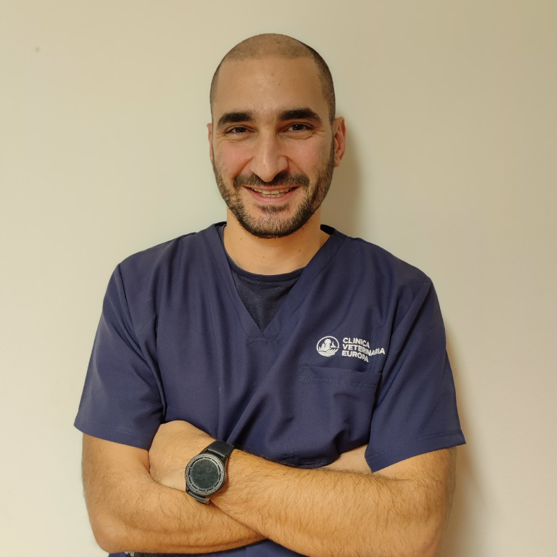 Un uomo con la testa e la barba rasate sorride con le braccia incrociate, indossa un camice da medico blu navy e un orologio nero, in piedi davanti a una parete chiara.