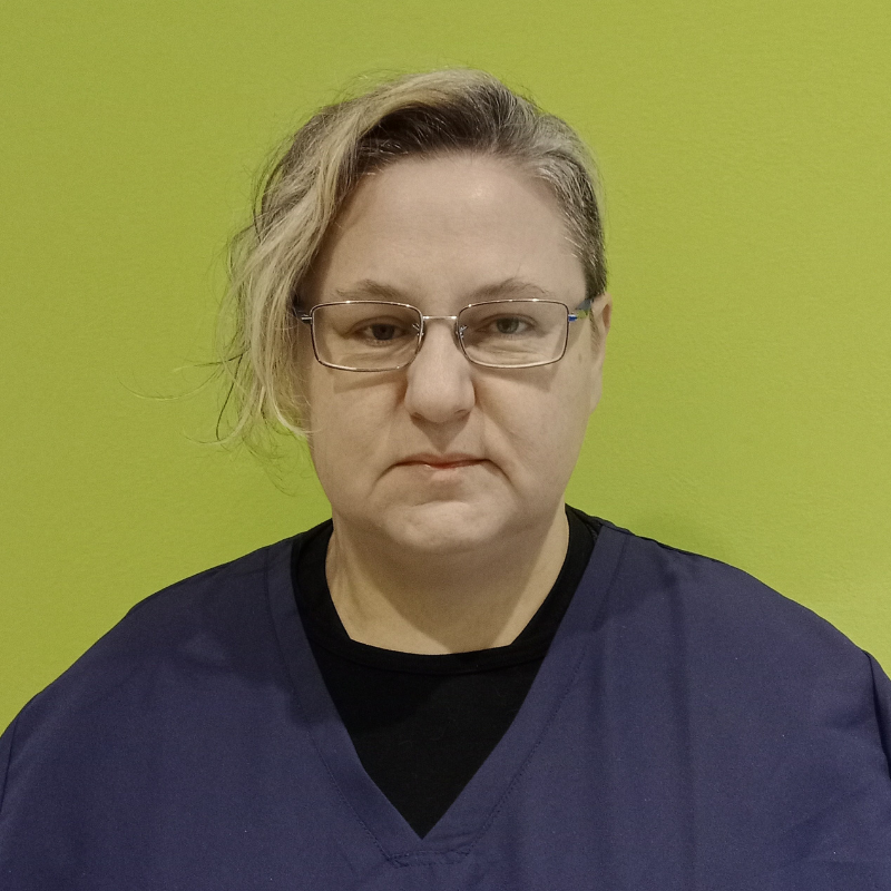 Una persona con capelli corti, castano chiaro e occhiali, che indossa un top blu navy, si trova su uno sfondo verde brillante e guarda direttamente la telecamera con un'espressione neutra.