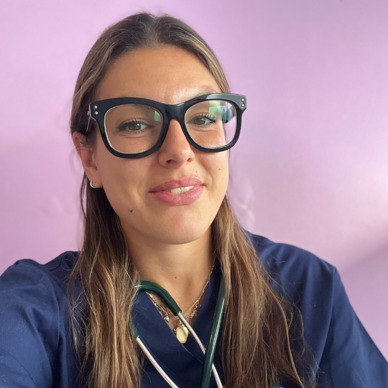 Una donna con lunghi capelli castani e occhiali neri, che indossa un camice blu e uno stetoscopio, sorride davanti a uno sfondo viola chiaro.