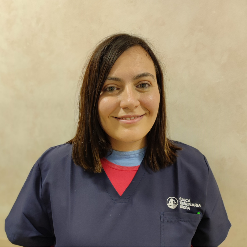 Una donna con i capelli lisci e castani che indossa un'uniforme blu navy con il logo della clinica veterinaria sorride alla telecamera su uno sfondo beige.