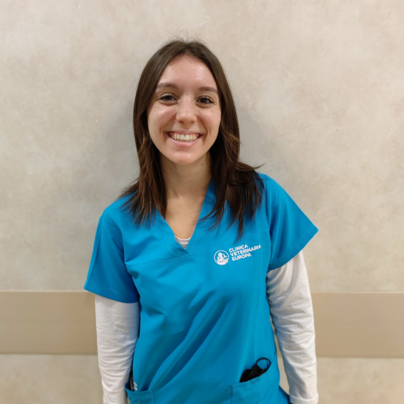 Una giovane donna con i capelli lisci e castani, che indossa un camice da medico blu su una camicia bianca a maniche lunghe, sta in piedi sorridendo contro una parete beige.