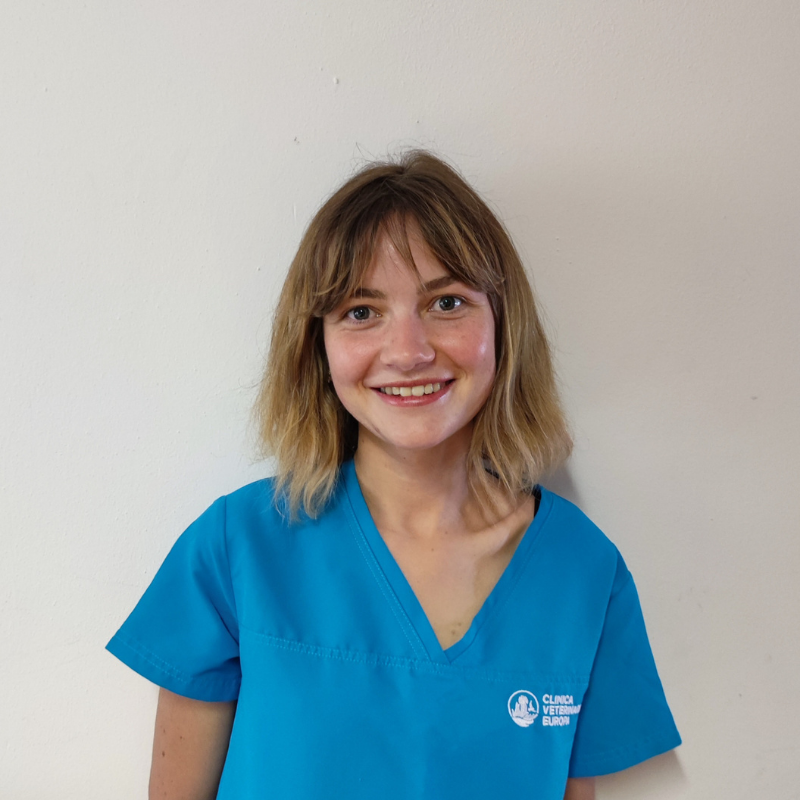 Una giovane donna con i capelli biondi lunghi fino alle spalle sorride alla telecamera. Indossa un camice medico blu con il logo bianco della clinica e si trova in piedi contro una parete bianca.