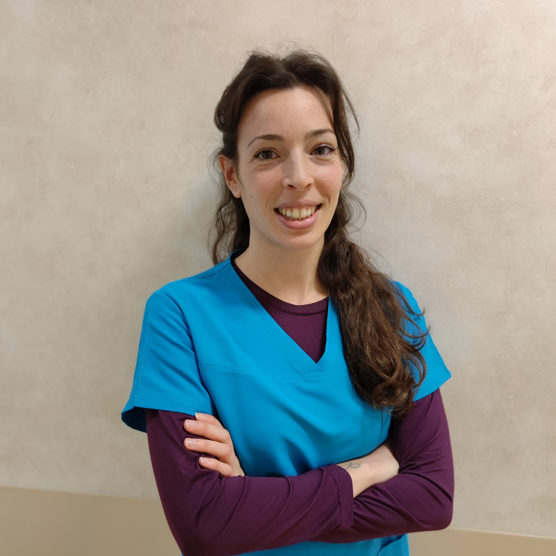 Una donna dai lunghi capelli castani, che indossa un camice da medico blu su una camicia viola, sta sorridendo con le braccia incrociate davanti a una parete chiara.