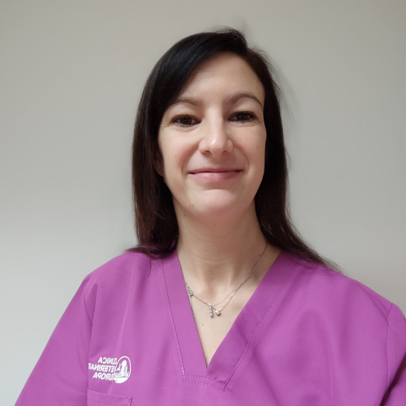 Una donna con i capelli lisci e scuri, che indossa un camice rosa e una collana, sorride alla telecamera su uno sfondo chiaro. Sul camice c'è il logo di una clinica veterinaria.
