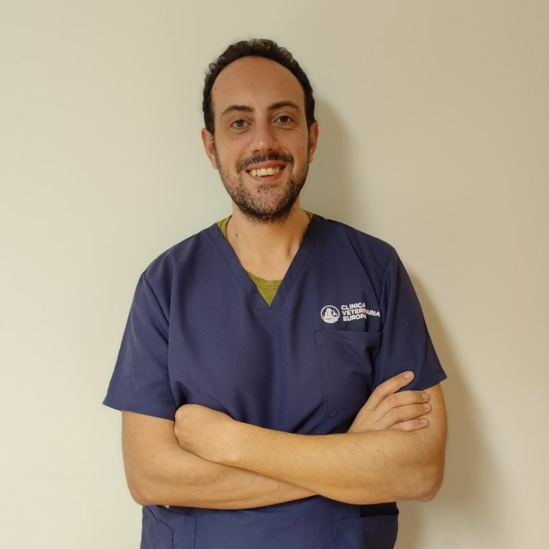 Un uomo con capelli corti e ricci e barba, che indossa un camice da medico blu navy, se ne sta in piedi sorridente con le braccia incrociate, pronto a sostenere Il team di fronte a un semplice muro chiaro.