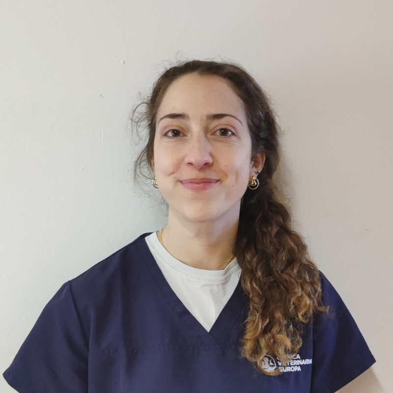 Una donna con lunghi capelli ricci e castani, che indossa un camice blu navy su una camicia bianca, è in piedi contro una parete chiara e sorride alla telecamera, rappresentando con orgoglio la squadra.