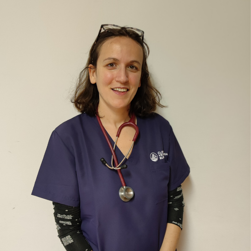 Una donna con un camice da medico viola e uno stetoscopio al collo sorride alla telecamera, rappresentando il team. Ha gli occhiali appoggiati sulla testa e si trova su uno sfondo chiaro.